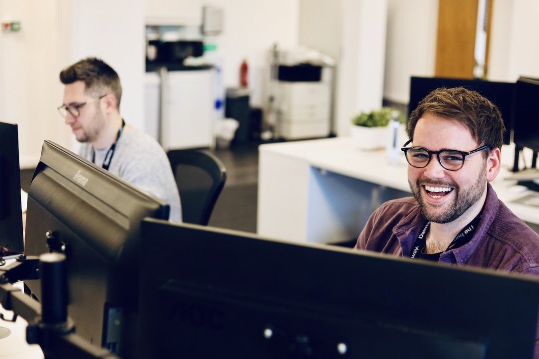 Two web designers working at their desks in a busy office, with one designer looking at the camera while smiling.