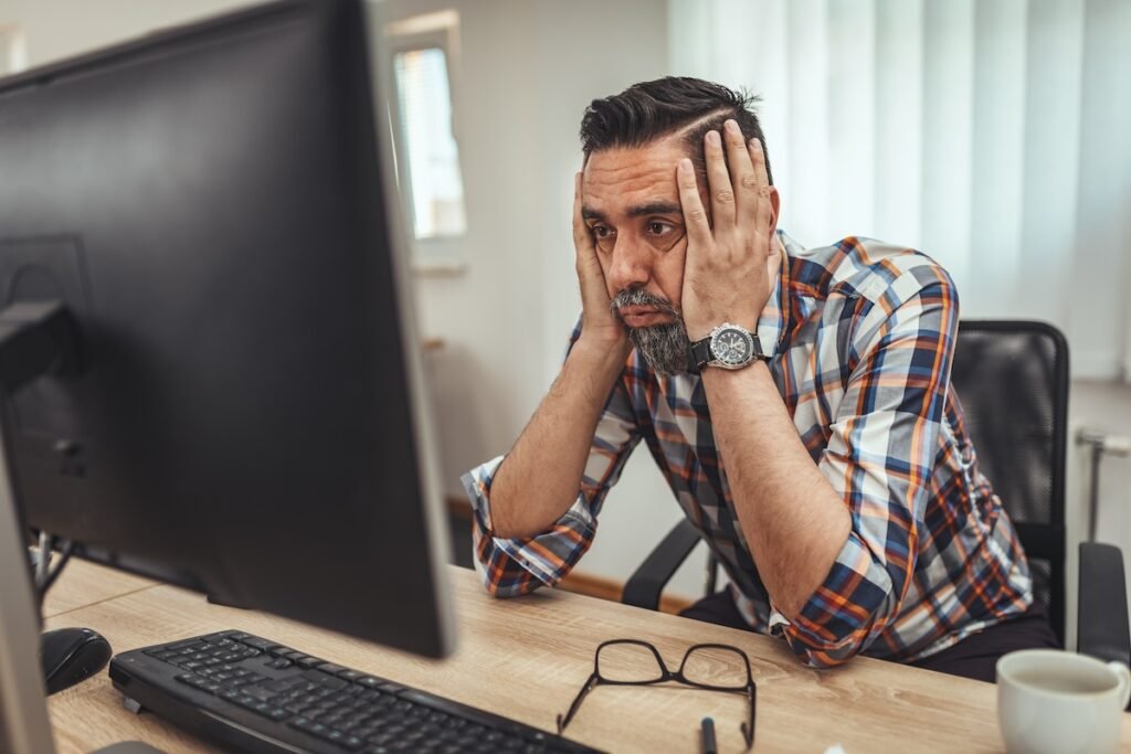 A businessman getting frustrated with a confusing and poorly structured website. He's sat a his desk, holding his head in his hands whilst staring at the computer monitor.
