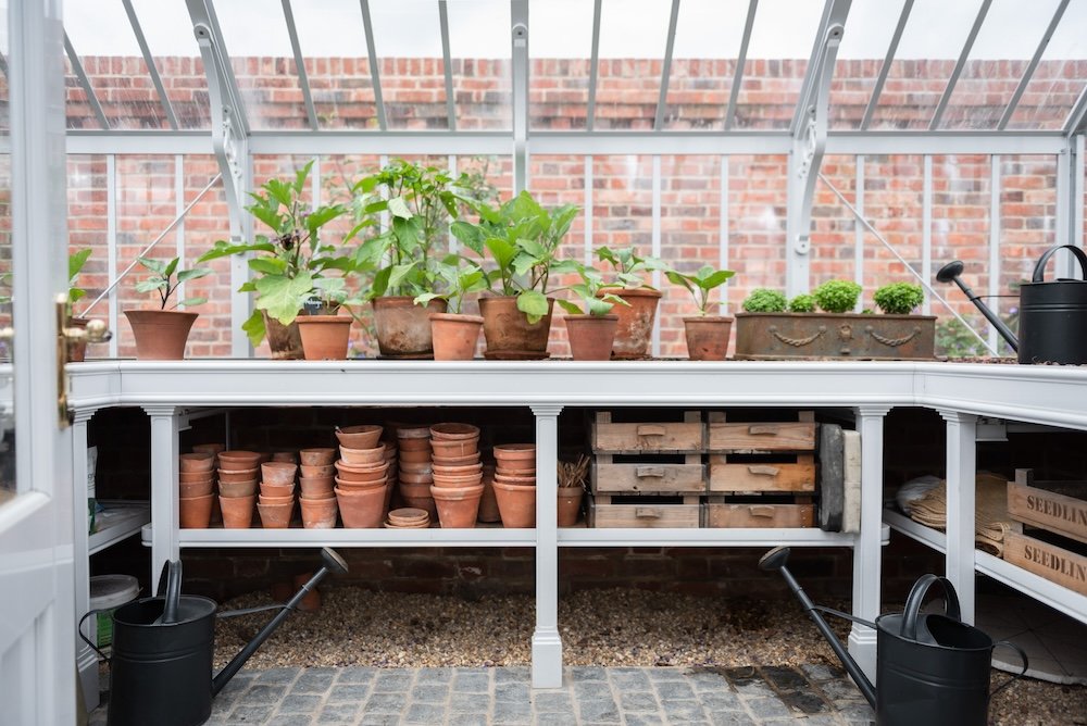 The inside of an Alitex greenhouse with a variety of clay plant pots, watering cans, and a gravel flooring.