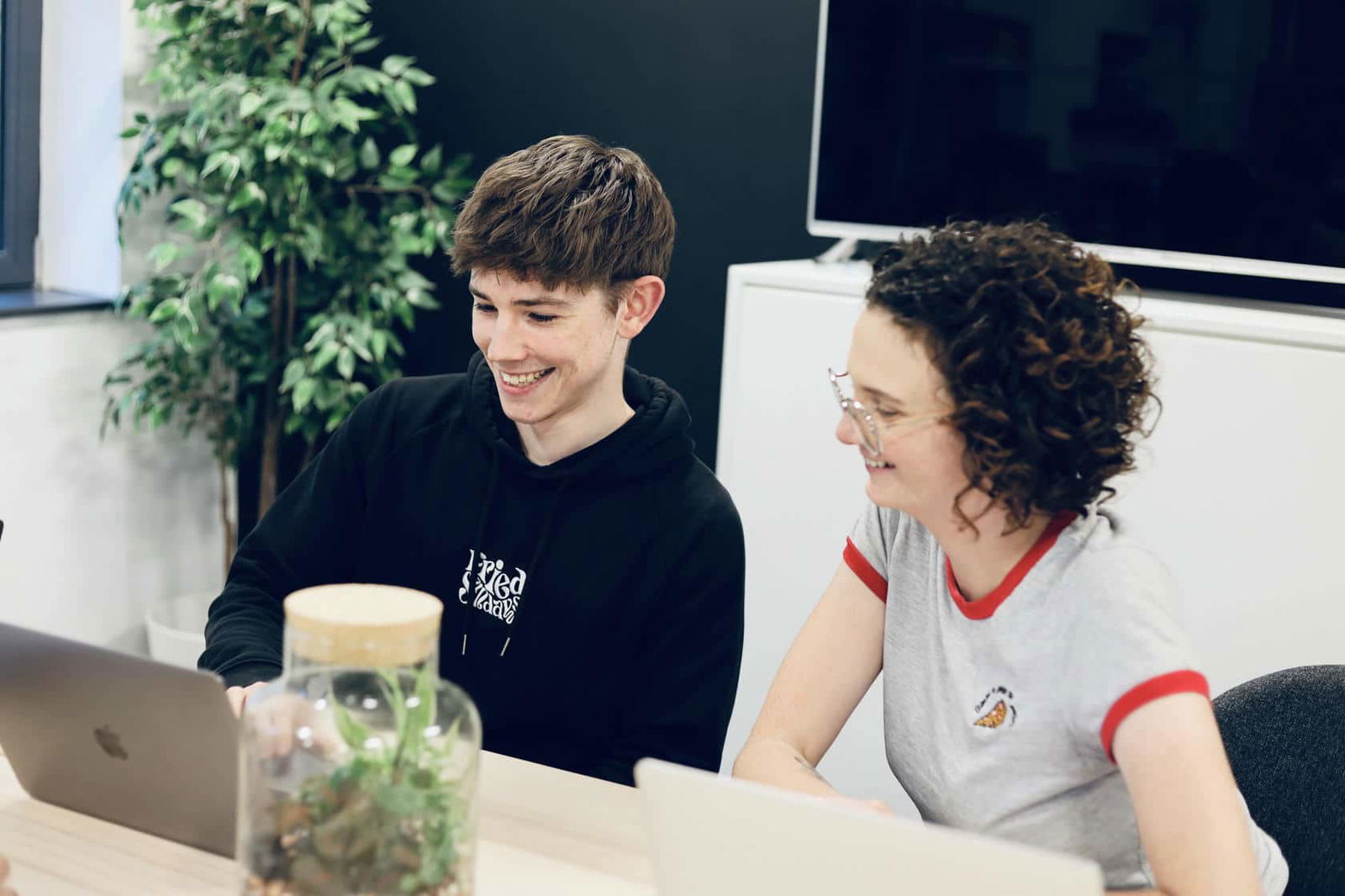 Ed and Emily, Damteq's UX specialists working smiling and working together on their laptops in a bright office.